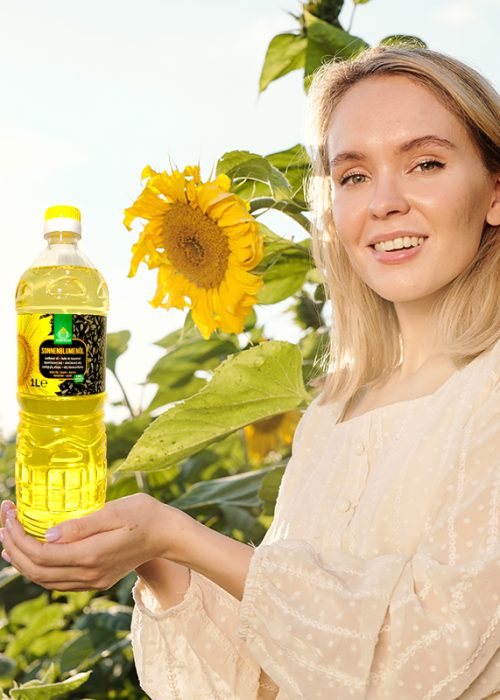 Smiling blond young woman in white dress holding bottle of sunflower oil while standing against large flowers in front of camera in the field